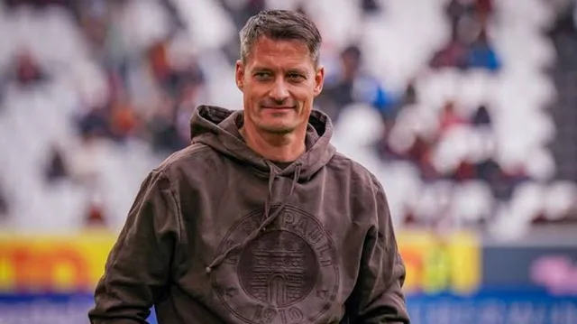 Alexander Blessin, kahverengi kapüşonlu sweatshirt ile stadyumda gülümseyerek yürüyor