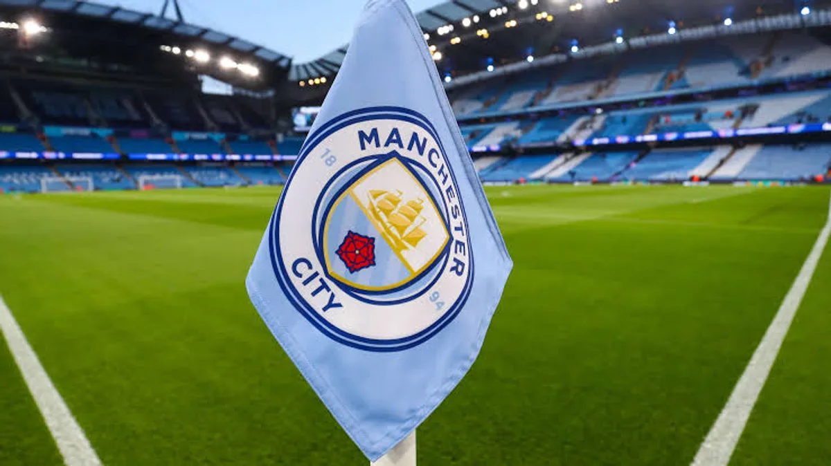 Manchester City armalı açık mavi korner bayrağı, arkada tribünleri ve çim sahasıyla stadyum